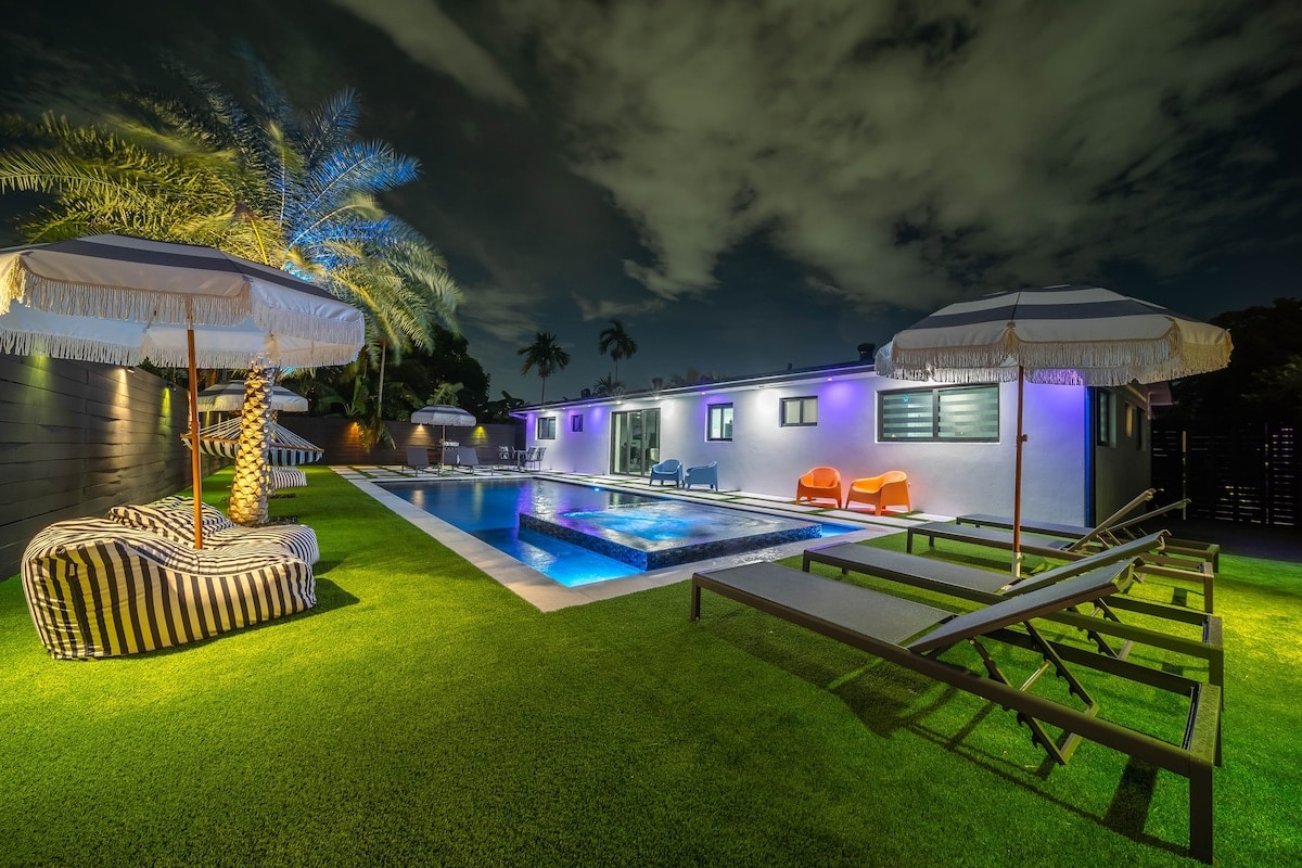 An inviting outdoor space features a pool surrounded by lounge chairs and umbrellas. Colorful seating is positioned along a well-manicured lawn, with palm trees providing shade. The structure is illuminated gently, creating a serene evening atmosphere.
