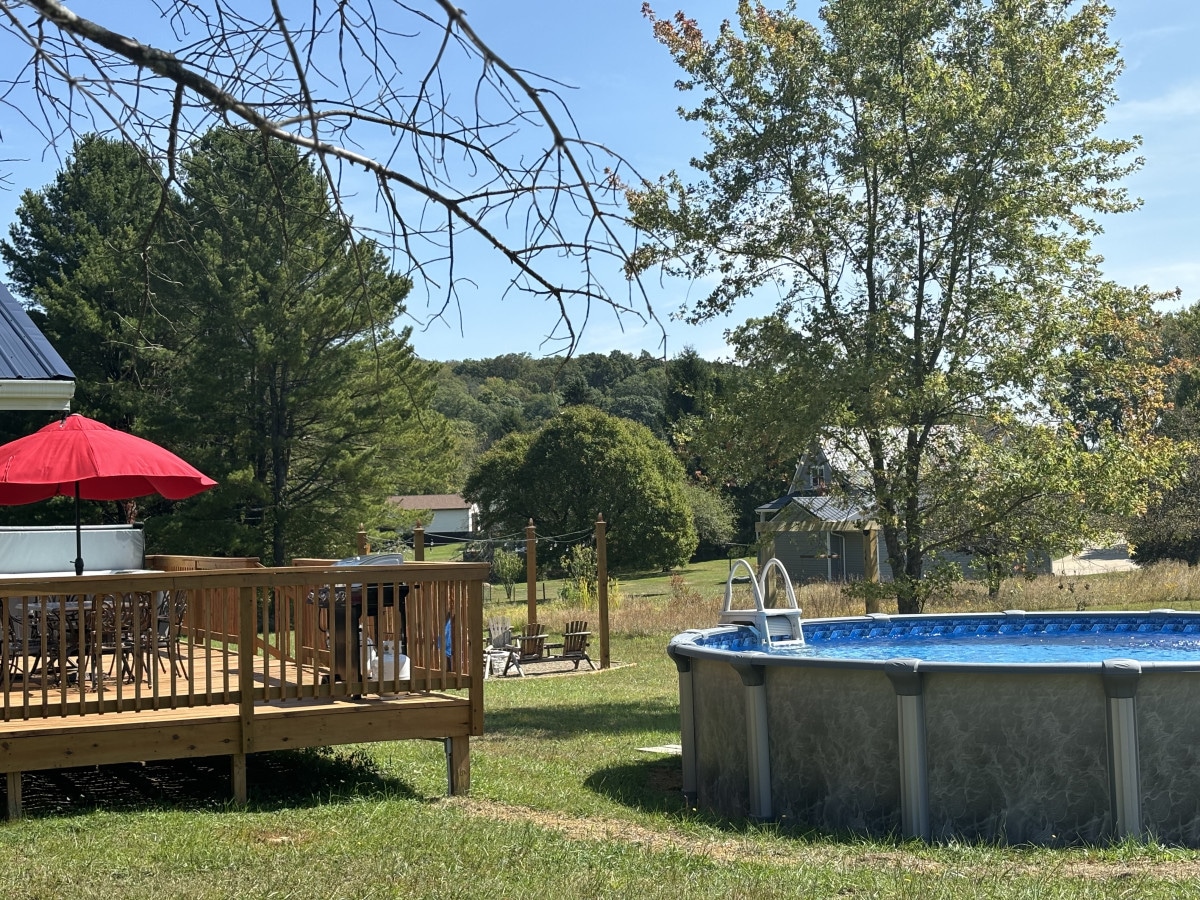 A family-sized pool is positioned beside a spacious deck, showcasing an umbrella-covered dining area. Lush greenery surrounds the scene, with trees and a distant structure visible in the background, providing a serene outdoor environment.