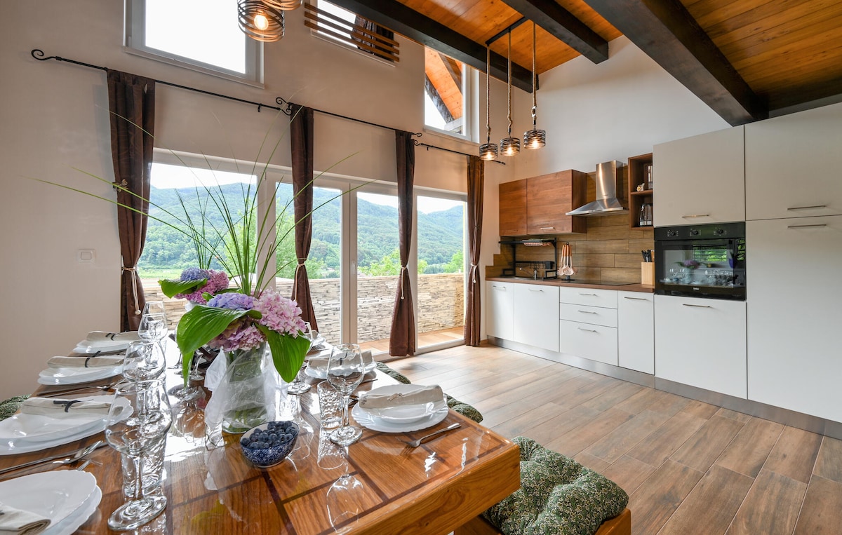 A dining area features a large wooden table set with white plates and glassware, accompanied by a decorative centerpiece of flowers. Large windows allow natural light to illuminate the space, revealing views of the surrounding landscape. Modern kitchen appliances are visible in the background.