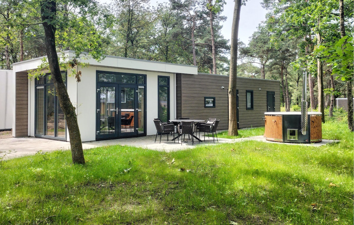 The holiday home is situated in a serene forest environment, featuring a modern exterior with large windows. A circular wooden bath is positioned near a dining table with chairs, surrounded by lush green grass and trees.