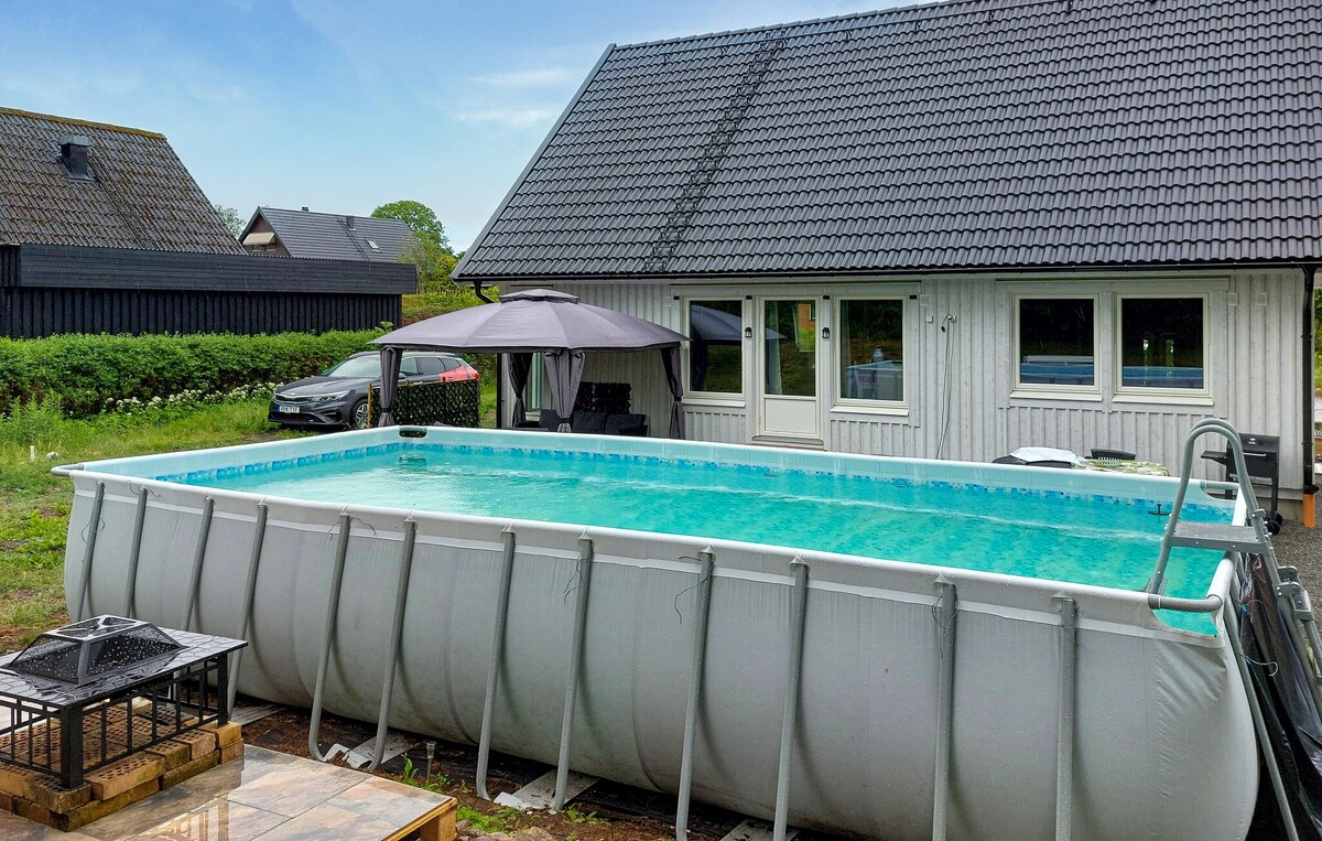 A heated outdoor pool is positioned in the yard, offering a refreshing blue surface. A modern white house with a gray roof is visible in the background, accompanied by a shaded seating area under a canopy. Lush greenery surrounds the property.