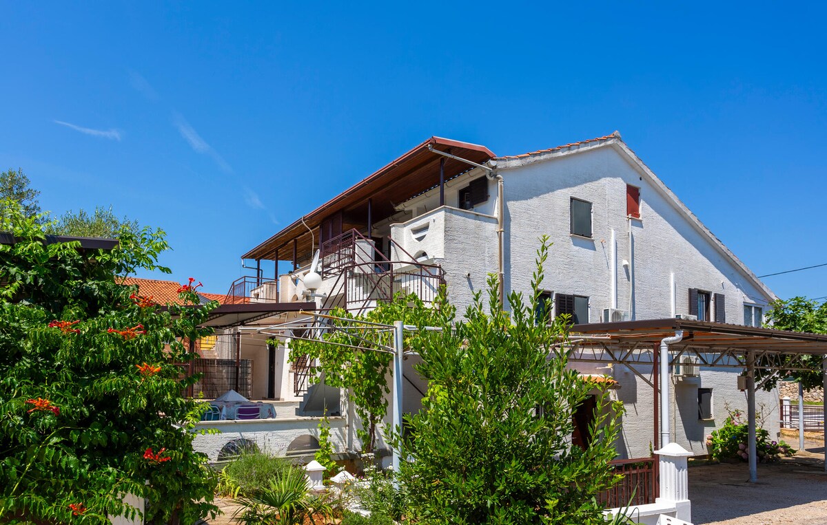 The exterior of the apartment building is presented against a clear blue sky. The structure is characterized by multiple balconies, surrounded by greenery and colorful flowers. A shaded terrace area is visible, providing a space for outdoor relaxation or dining. A pool area is also included in the landscape.