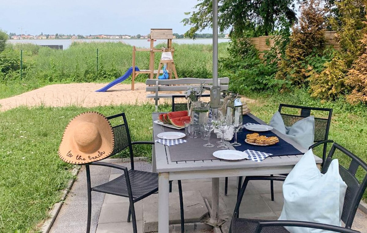 An outdoor dining area is presented with a wooden table set for a meal, complemented by black chairs. A large sunhat rests on the table. In the background, a playground and open grassy space are visible, providing a view of the lake beyond.