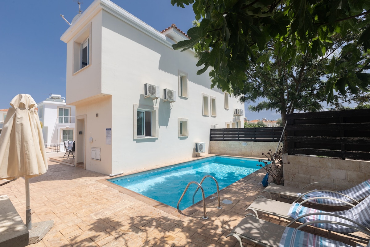 The exterior view of the villa showcases a private pool surrounded by a tiled patio. Sun loungers are positioned by the pool, and a parasol offers shade. The white building features several windows and air conditioning units, framed by greenery and a wooden fence.