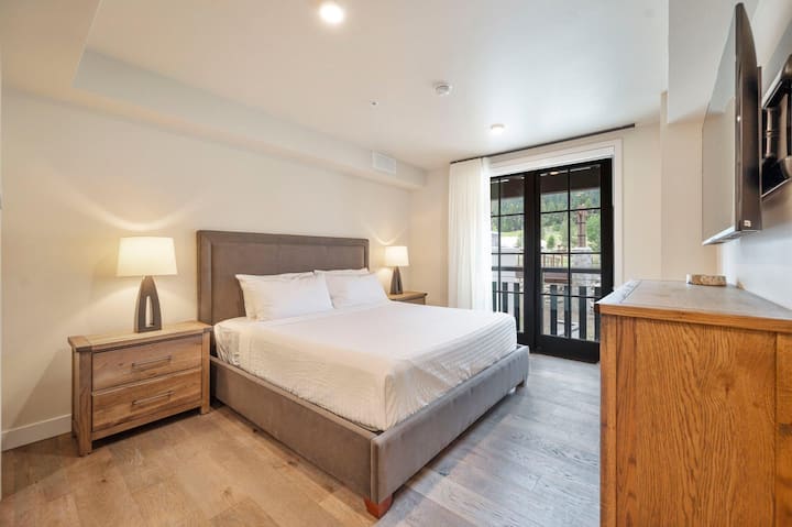Crisp white linens and a leather-paneled headboard set a restful tone in the upper-level king suite, with a weathered-oak dresser, wall-mounted TV, and balcony access framing green mountain views beyond