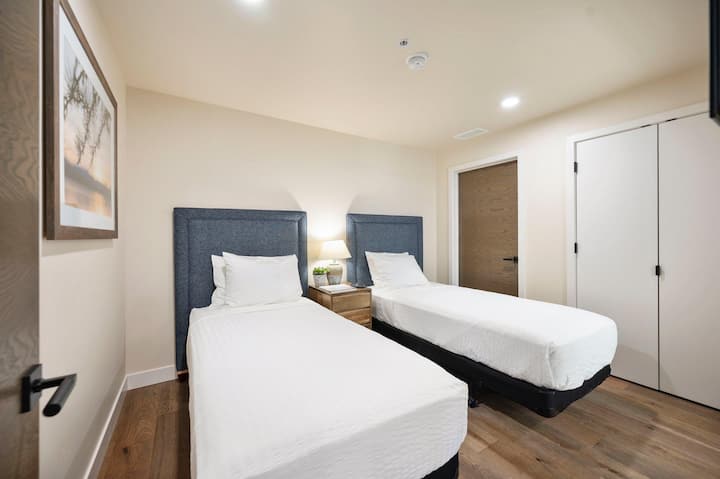 Two twin beds with navy upholstered headboards share a warm bedside lamp and framed nature art — a cozy upper-level room with plenty of closet space for young adventurers or extra guests at Tamarack.
