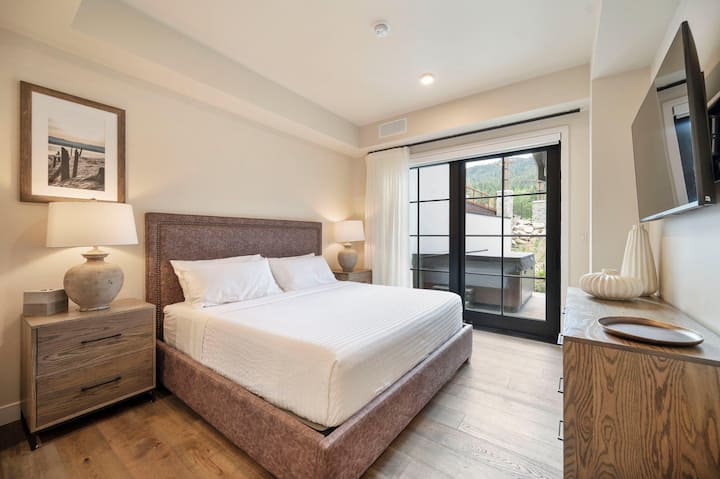 The main-level king suite features a nailhead-trimmed headboard, ceramic bedside lamps, and sliding glass doors that open directly to the private patio and hot tub — mountain views waiting just outside.