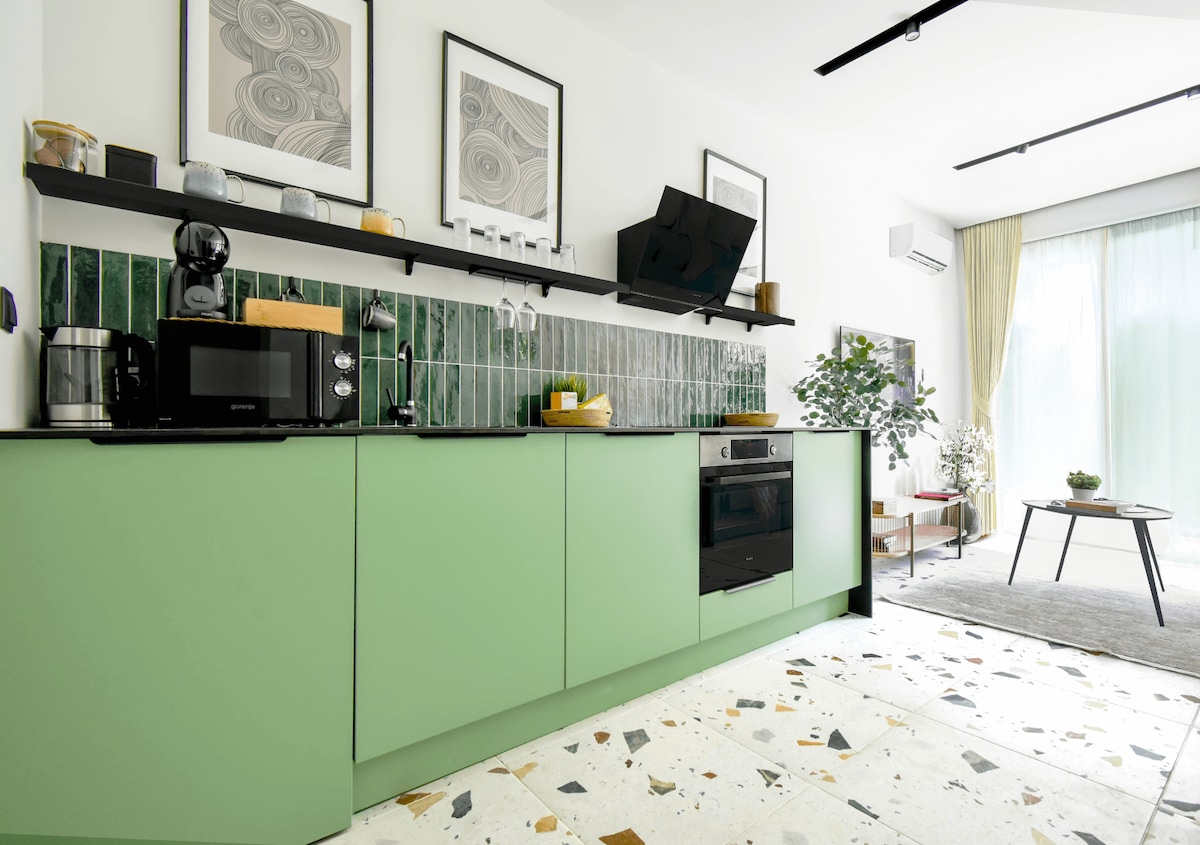A modern kitchenette showcases a refreshing green cabinet and a sleek oven. Essential appliances, including a coffee machine and microwave, are neatly arranged on the countertop. Artwork adorns the walls, and natural light streams in through large windows, illuminating the space.