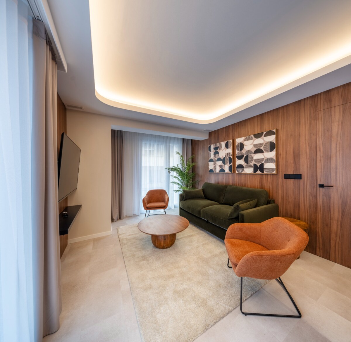 A modern living area features a large green sofa complemented by two accent chairs in warm tones. A round wooden coffee table is placed atop a light-colored area rug. The space is illuminated by soft ceiling lights, with large windows draped in sheer curtains.