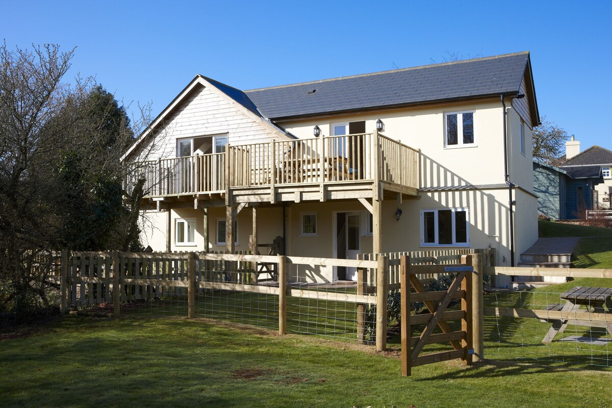 The exterior of the house showcases a light-colored facade with a spacious balcony adorned with wooden railings. The landscaped yard is enclosed by a wooden fence, featuring a gate that leads to the entrance. Surrounding greenery offers a natural backdrop.