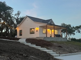 A newly-built farmhouse cottage is nestled among oak trees, showcasing a light-colored exterior with windows that reflect the evening glow. A concrete path leads to a covered porch, where seating is positioned for relaxation amidst the serene country setting.