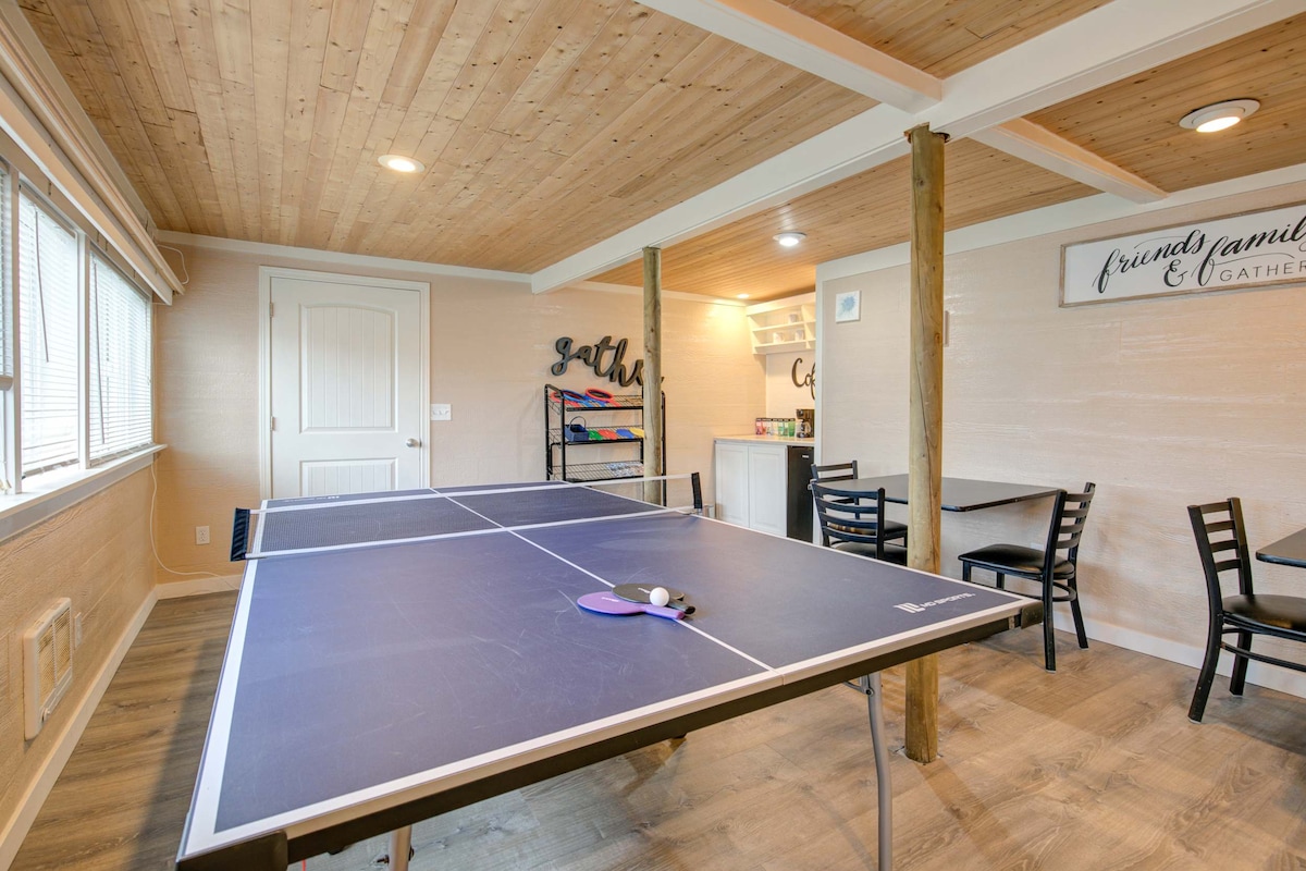 A recreational space features a ping pong table centrally located, with chairs arranged around a dining area in the background. Natural light enters through large windows, illuminating the warm wooden ceiling and walls, creating a welcoming environment for social gatherings.
