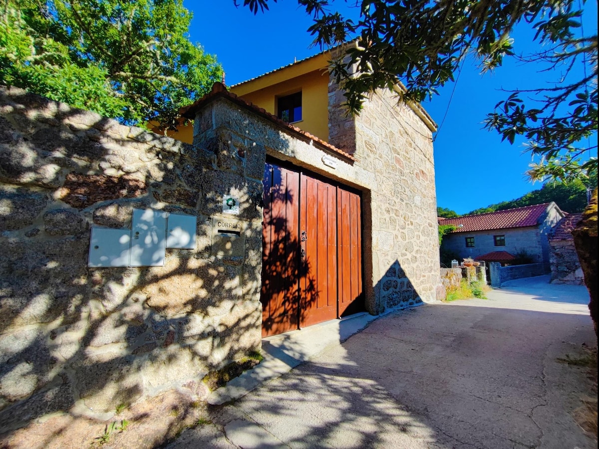 A stone wall surrounds a rustic wooden gate, marking the entrance to the property. A gravel path leads into the yard, embraced by trees and sunlight. In the background, the warm colors of the house and surrounding buildings complement the natural landscape.