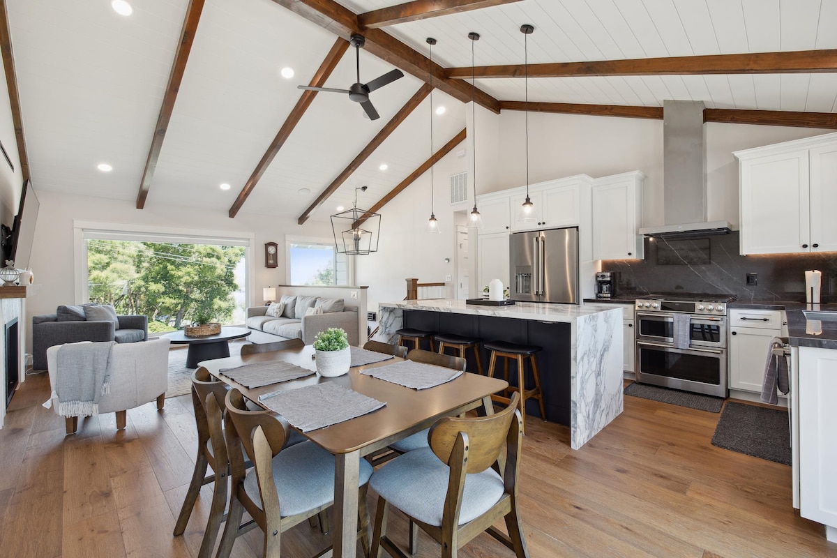 An open-concept living space is highlighted, featuring a modern kitchen with stainless steel appliances and marble countertops. A dining table set for six complements the casual seating area, adorned with comfortable sofas. Large windows allow natural light to illuminate the wood-beamed ceiling and stylish decor.