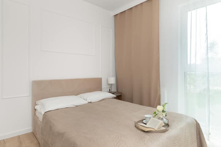 Удобна спална соба со неутрална постелнина и природна светлина што струи низ прозорецот промовира одмор и закрепнување.