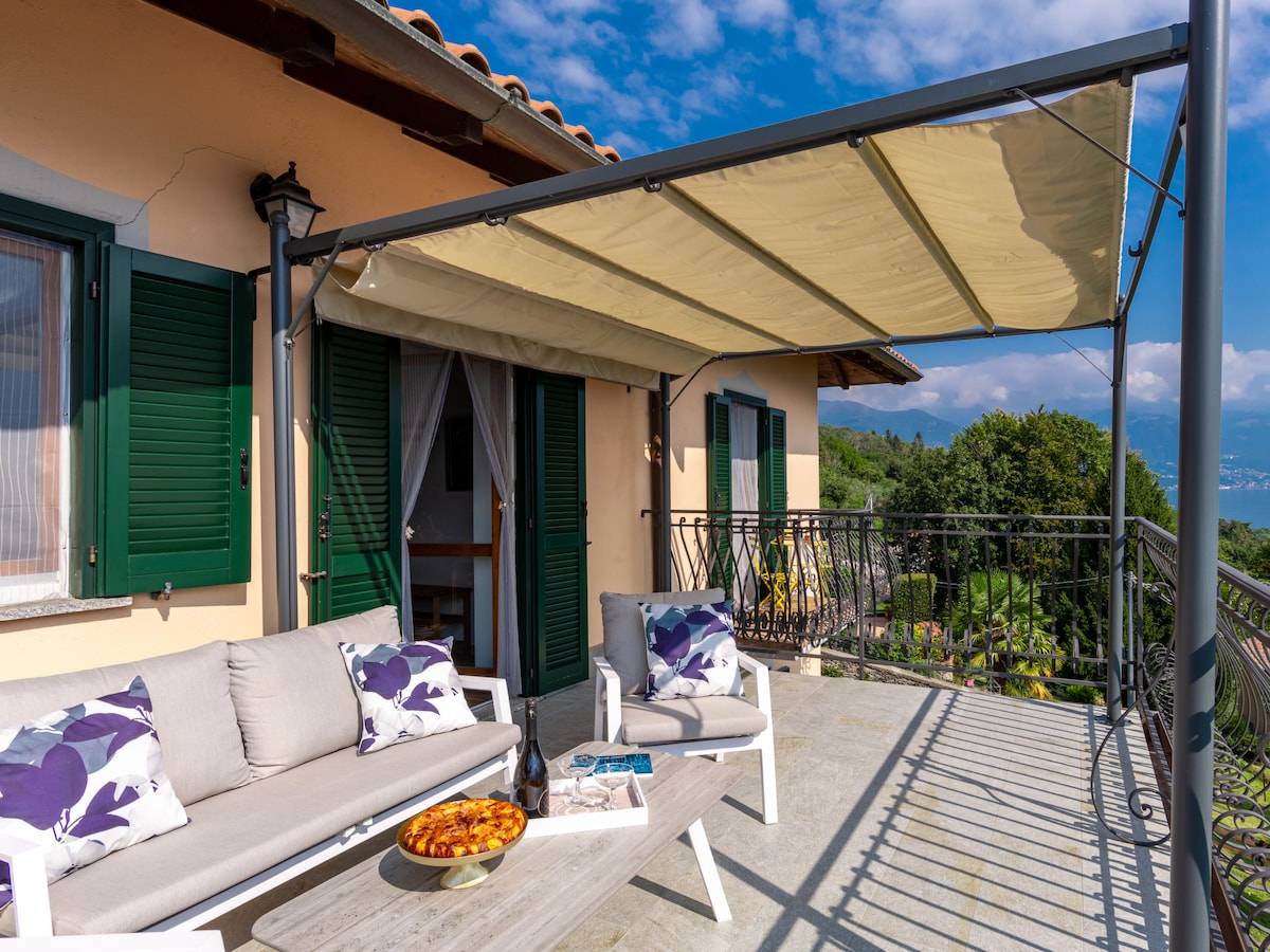 A spacious terrace features comfortable seating with cushions, complemented by a central coffee table. An awning provides shade over the area, which is surrounded by green trees and offers panoramic views of the landscape.