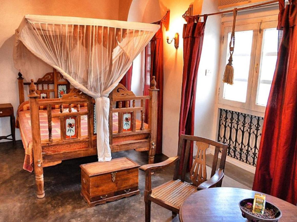 A double bed is framed with wooden furniture, featuring a canopy draped with sheer fabric. A small wooden storage chest is positioned at the foot of the bed. A round table and a wooden chair are set near the window, with red curtains providing warmth to the decor.