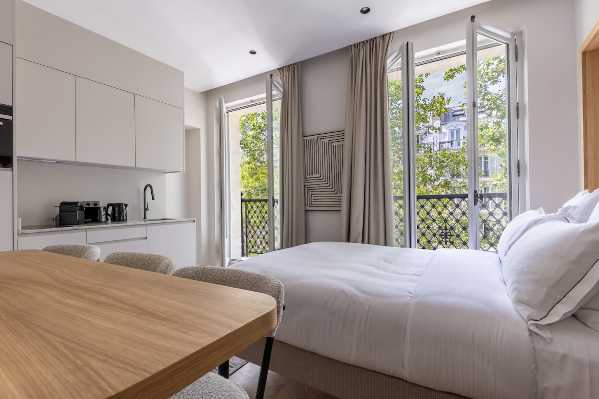 A modern studio features large windows that allow natural light to fill the room, framed by elegant curtains. A spacious bed is positioned across from a fully equipped kitchen area, with a dining table and chairs nearby. A calming artwork piece is displayed on the wall.