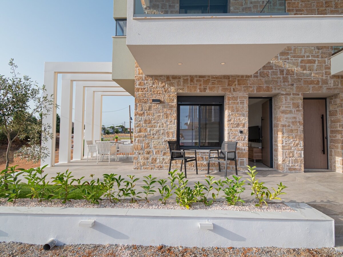 A modern outdoor patio area is visible, featuring a table and chairs, ideal for relaxation. The stone façade of the building provides a natural aesthetic. Lush greenery borders the space, with a clear view of the surrounding landscape and bright sky in the background.