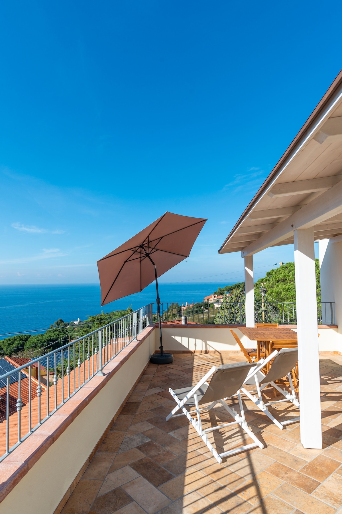 An outdoor terrace is shown, furnished with a wooden dining table and several chairs. A large umbrella provides shade while the expansive sea view stretches across the horizon under a clear blue sky.