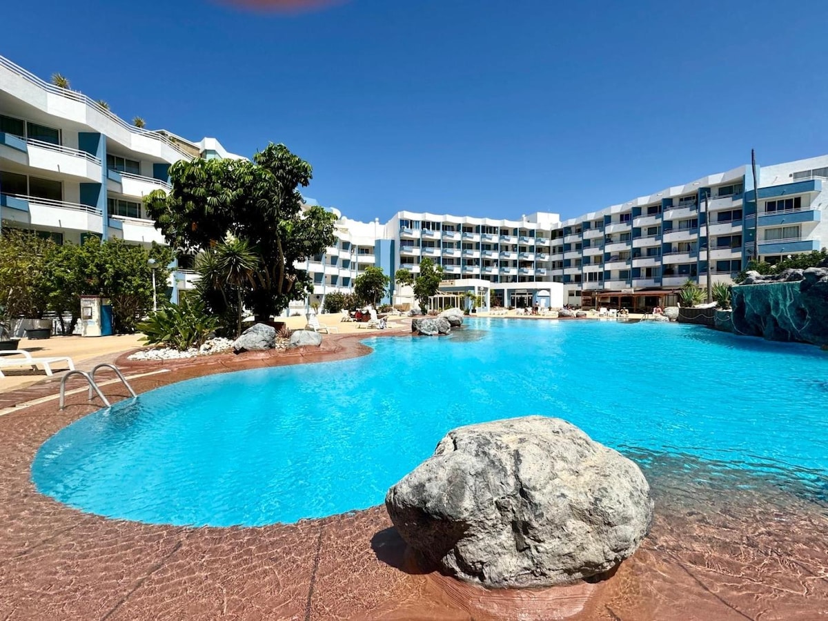 An expansive outdoor pool is framed by natural rock formations and tropical landscaping. Lounge chairs are arranged around the pool area, with bright blue water reflecting the clear sky. A modern apartment building in the background offers a contemporary architectural design.