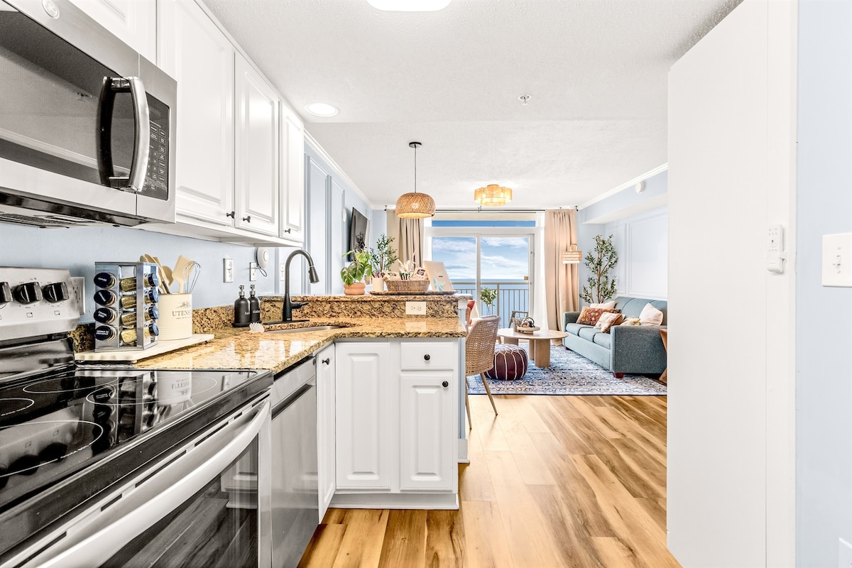 A modern kitchen is equipped with dark appliances, granite countertops, and white cabinetry. An open layout provides a clear view of the living area, where large windows frame ocean views. A cozy seating arrangement includes a neutral-colored sofa and decorative accents.