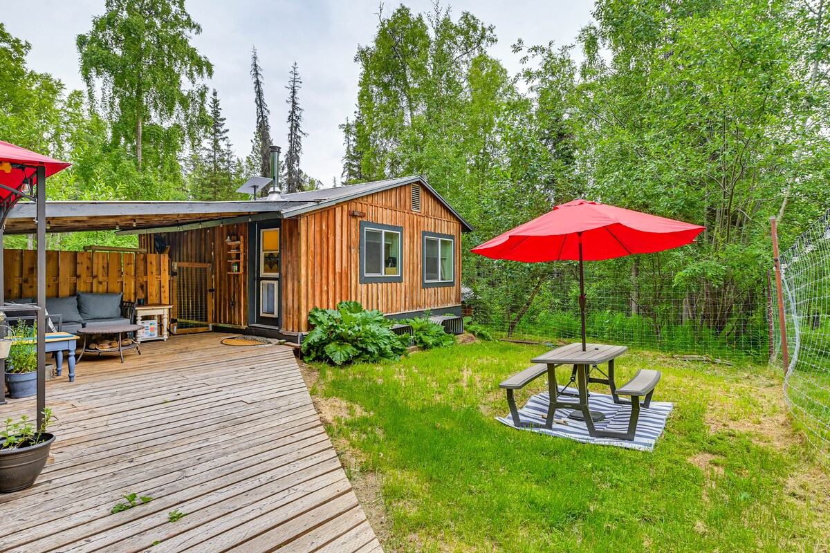 An outdoor living space showcases a wooden deck with seating areas and a picnic table beneath a bright red umbrella. Surrounding greenery enhances the natural setting, while the cabin features multiple windows that invite light and views of the outdoors.