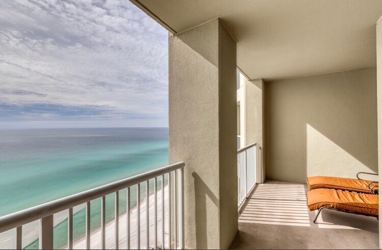 A private balcony is featured with a single lounge chair overlooking the Gulf of Mexico. The serene blue-green waters are visible, while soft clouds float in the sky. The balcony's railing offers unobstructed views of the beach below.