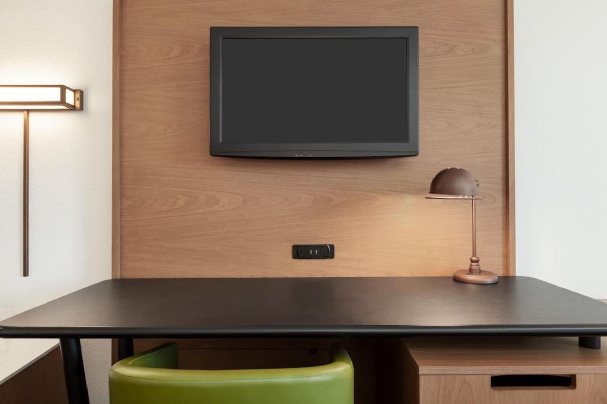 A functional workspace features a sleek black desk against a wooden wall. A green chair is positioned at the desk, complemented by a stylish lamp. Above, a flat-screen TV is mounted, providing entertainment options within reach.