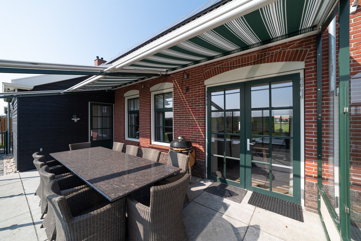 An expansive outdoor terrace is equipped with a large stone table, surrounded by comfortable rattan chairs, providing ample seating for ten. A striped awning offers shade, while large glass doors lead into the interior, highlighting the connection to the garden.