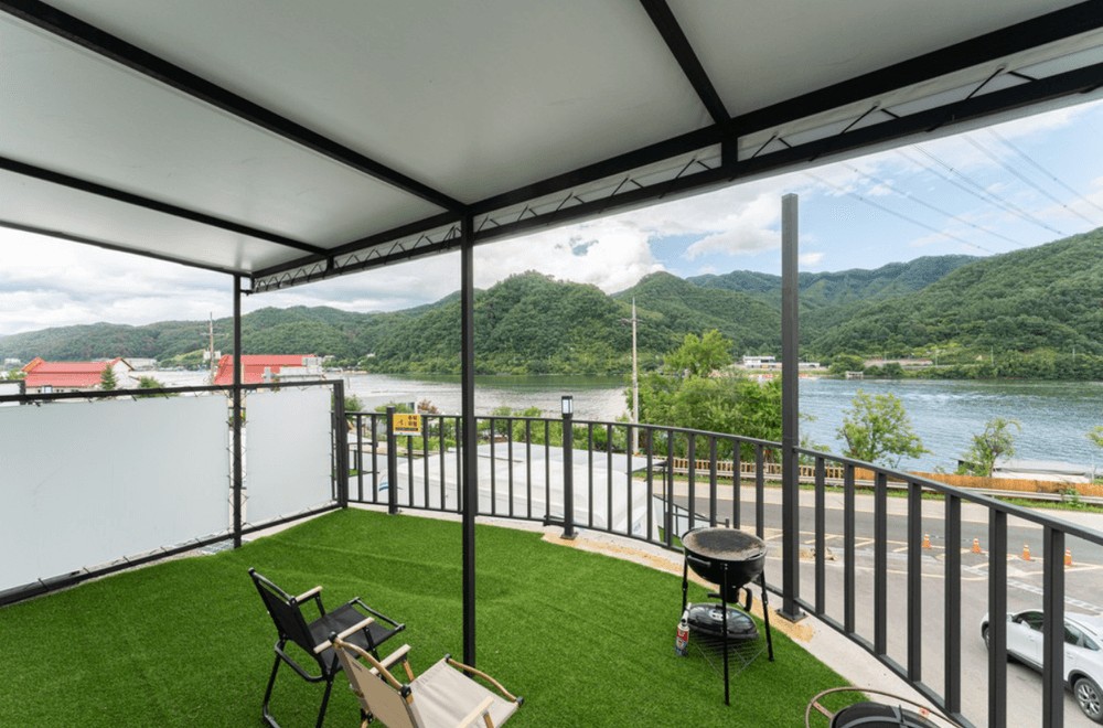 A spacious outdoor terrace features artificial grass, offering a comfortable seating area with two chairs. A barbecue grill is visible, and stunning views of the surrounding mountains and river are presented through large openings, creating an inviting space for relaxation.