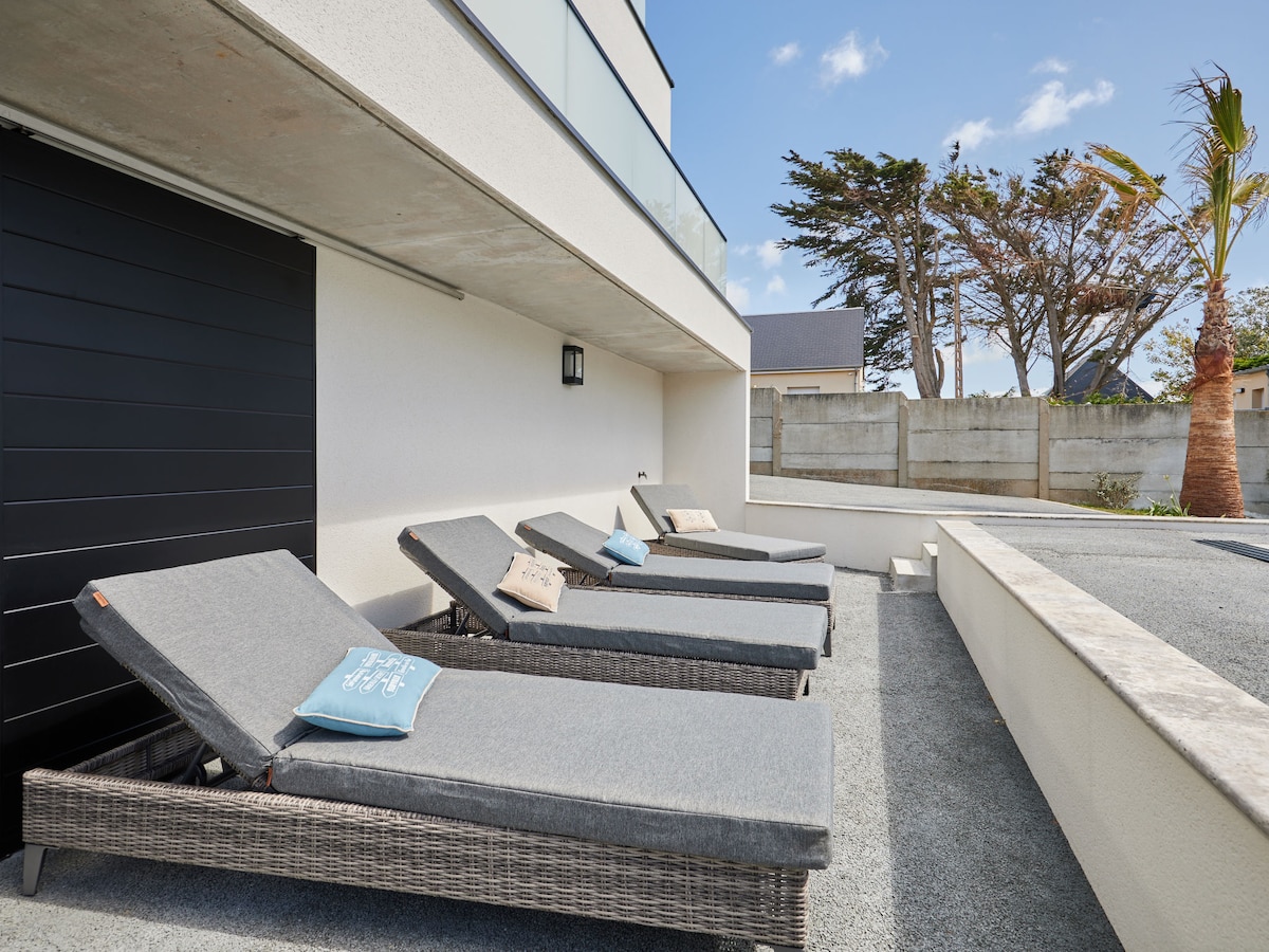 Four lounge chairs are arranged on a smooth outdoor surface, each with a light-colored cushion. The area is bordered by a lush garden and features a low wall. A clear sky is visible in the background, suggesting a sunny atmosphere.