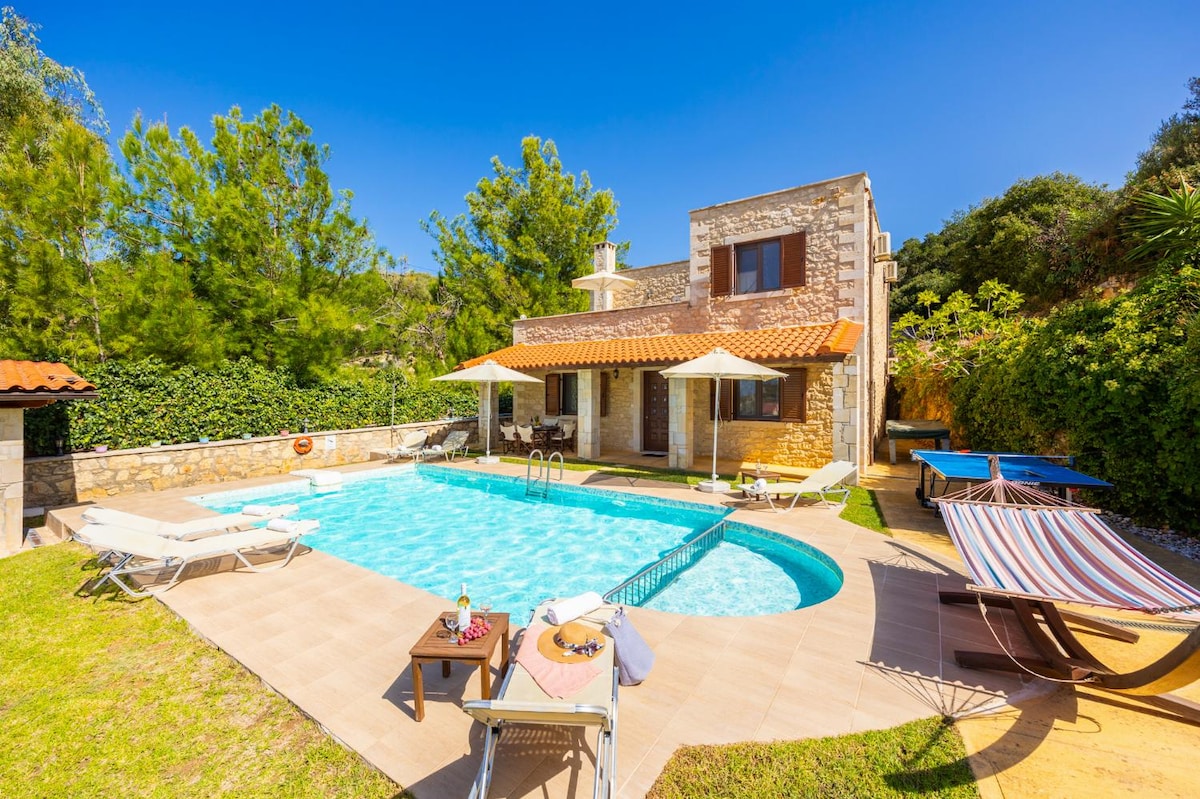 The private pool area features a rectangular pool with a shallow end. Surrounding the pool are sun loungers and a shaded dining table. A colorful hammock and additional seating can be seen to the side, with lush greenery and stone walls framing the scene.