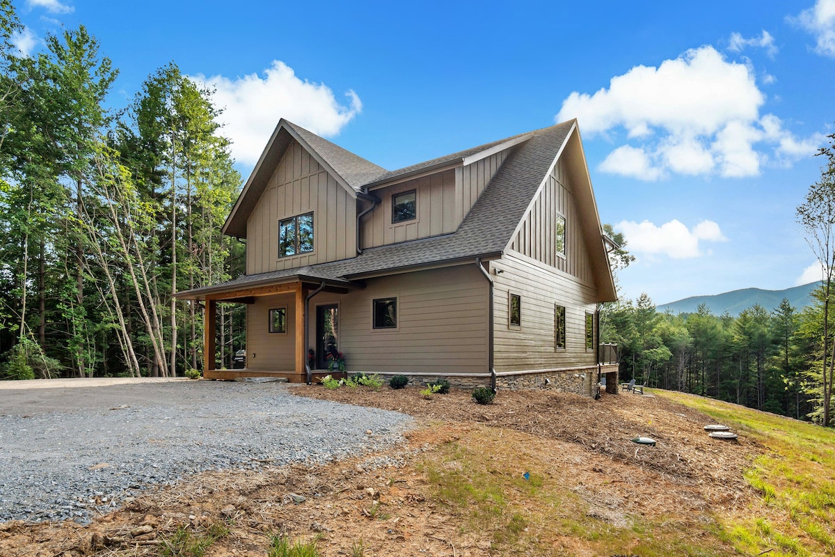 The exterior of a modern home is set within a natural landscape, featuring a combination of wood and stone finishes. Large windows allow for ample sunlight, and a stone pathway leads to the entrance. Surrounding trees provide privacy and a serene atmosphere.