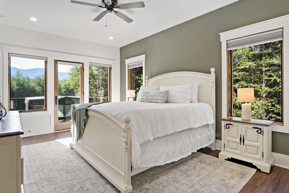 A spacious bedroom features a large, white bed dressed in light linens, complemented by side tables with lamps. Natural light floods through multiple windows, framing views of the surrounding greenery and mountains. A neutral area rug anchors the space.