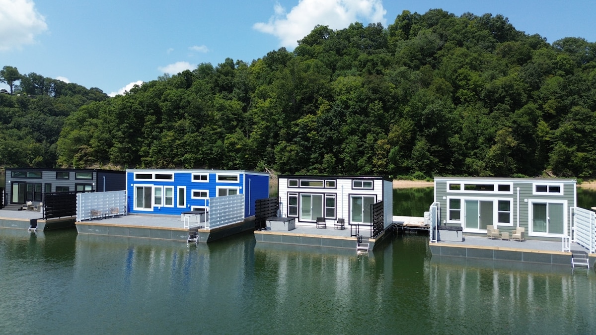 A cluster of modern float houses is shown anchored on calm waters. Each house features large windows for ample natural light and a blend of distinctive colors. Lush greenery lines the backdrop, enhancing the serene lake atmosphere.