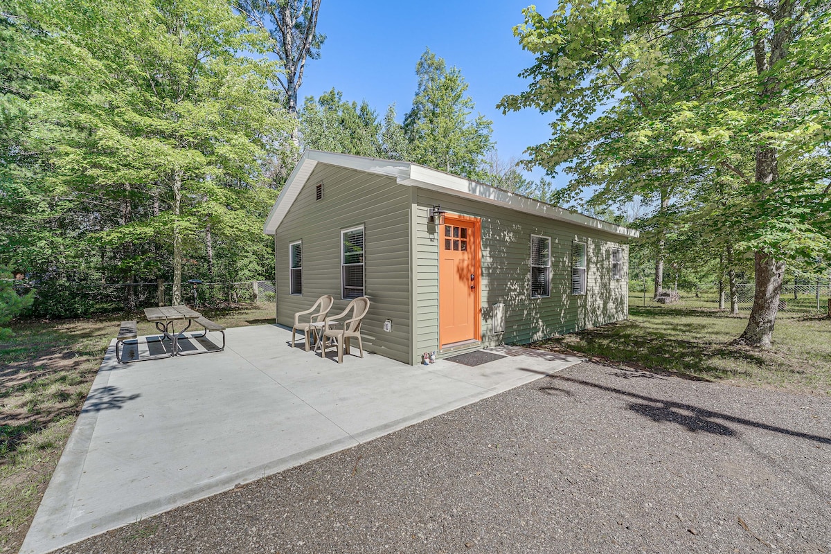 A cabin exterior is shown, featuring a bright orange door and several windows. A spacious concrete patio is visible, furnished with wooden picnic tables and plastic chairs. Surrounding trees provide a natural setting, enhancing the overall peaceful atmosphere of the property.