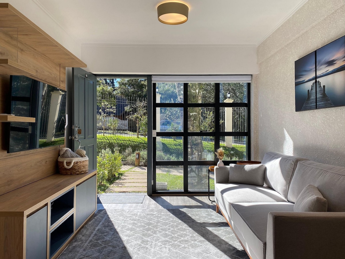 A well-lit living area features a comfortable gray sofa and a stylish wooden console with media storage. Large glass doors open to a garden view, allowing natural light to fill the space. A piece of art depicting a pier is displayed on the wall.