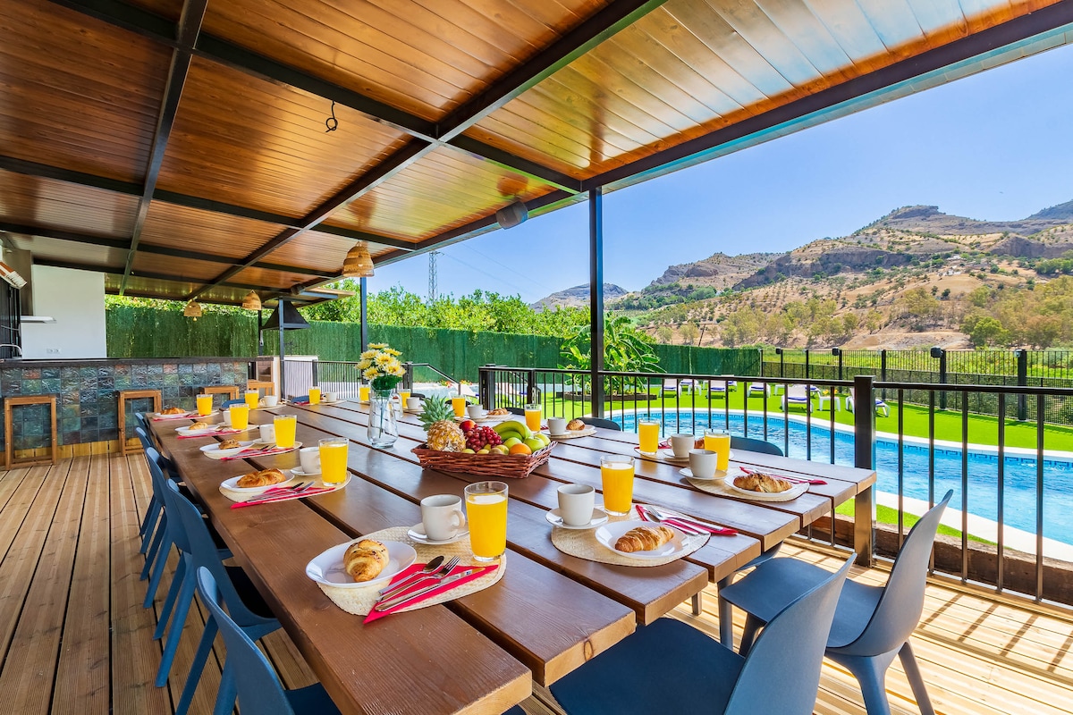 An outdoor dining area features a long, wooden table set for breakfast, with plates, croissants, and juice glasses arranged. A view of the private swimming pool and surrounding hills is visible, complementing the inviting space adorned with flowers and natural light.