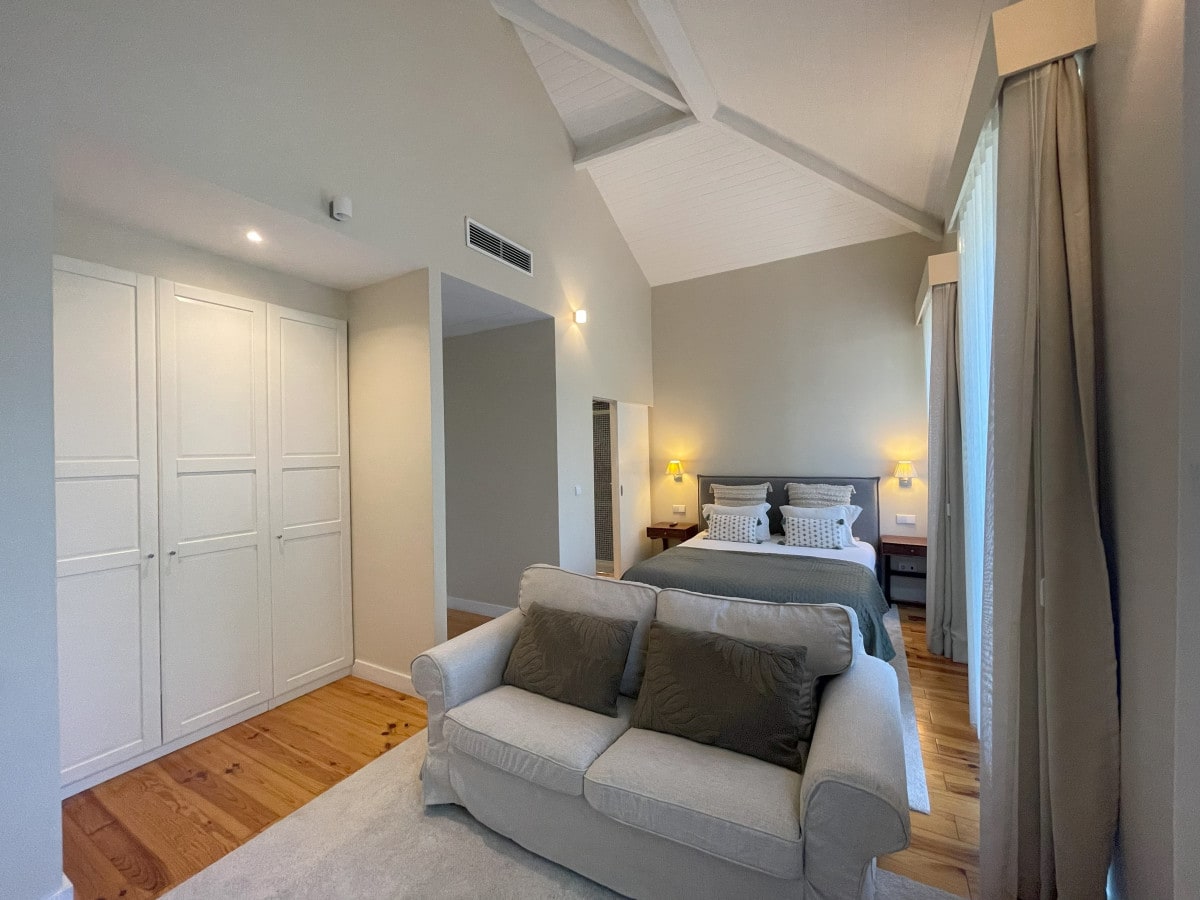 A spacious room features high ceilings with wooden beams, a comfortable bed with decorative pillows, and a cozy sofa. Natural light filters through large windows, illuminating the stylish wardrobe and bedside tables with lamps.