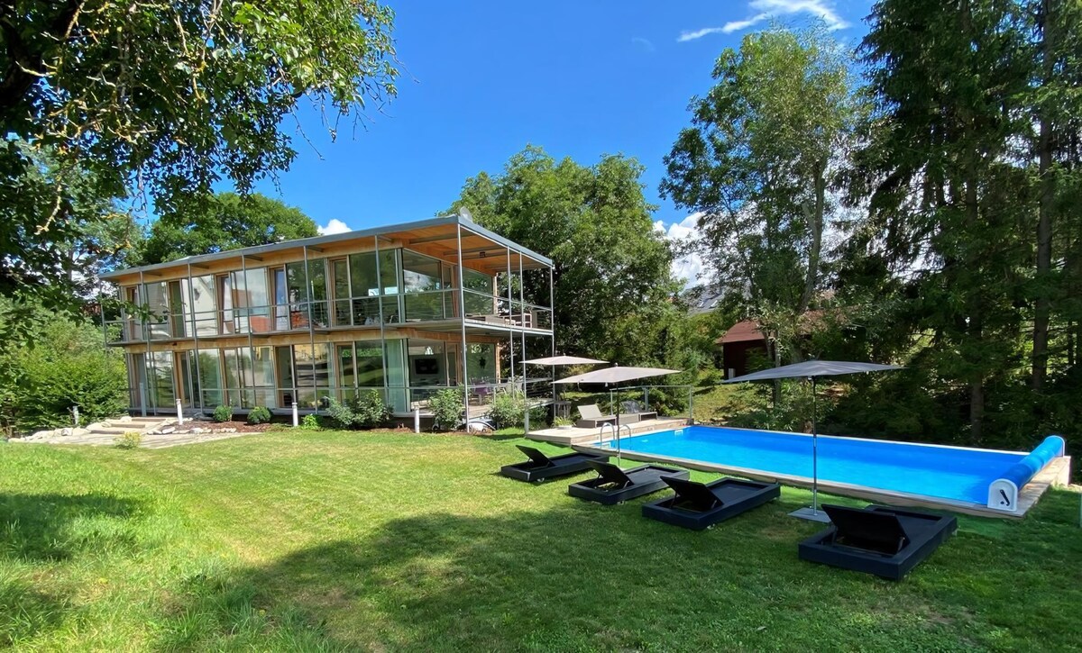 A modern two-story holiday home is framed by lush greenery and a blue sky. The spacious lawn features four lounge chairs and an outdoor infinity pool, highlighting the seamless connection between the indoor and outdoor living spaces.