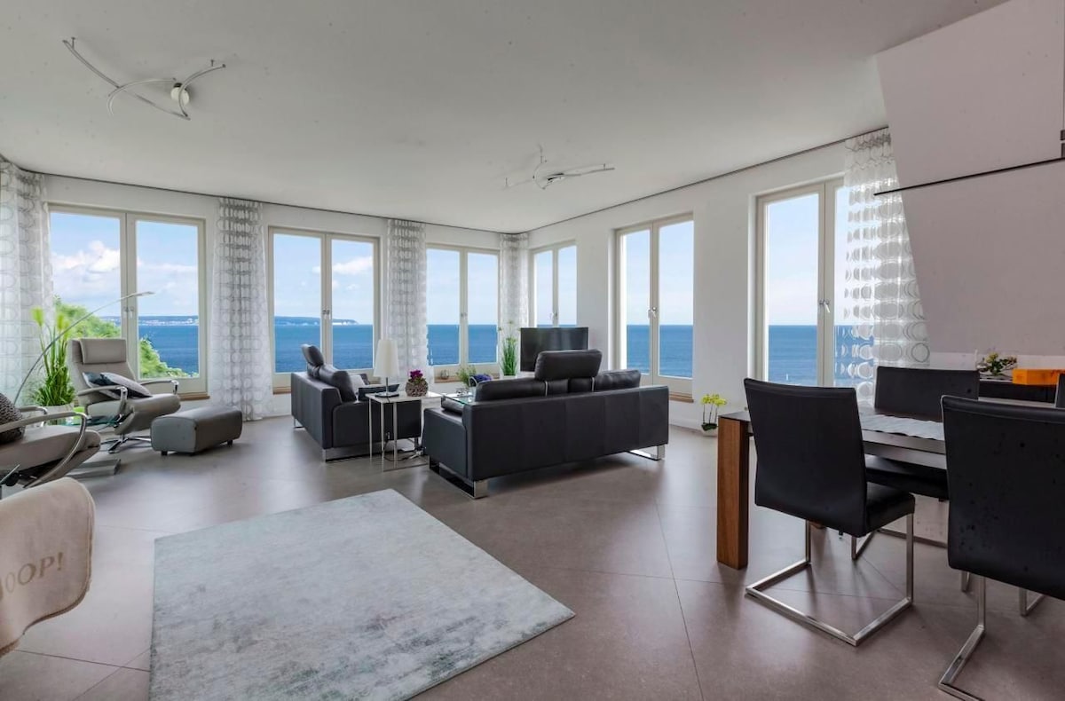 A modern living area features large windows that offer expansive sea views. Comfortable black sofas are positioned invitingly around a coffee table. A dining area with sleek furnishings is visible, while a light rug adds softness to the space. Natural light fills the room, enhancing a bright atmosphere.