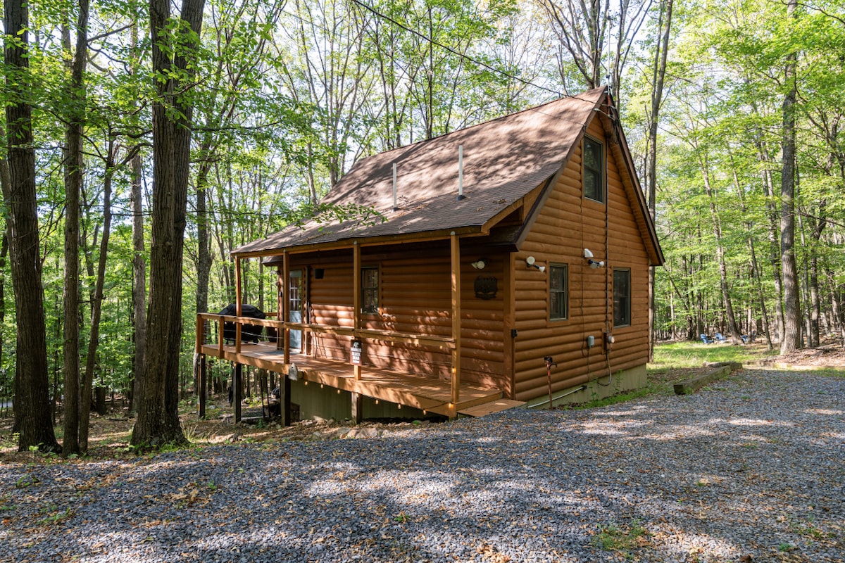 A wooden cabin nestled among trees is highlighted by a spacious deck, ideal for outdoor gatherings. The brown exterior blends harmoniously with its natural surroundings. Gravel pathways lead to the cabin, offering access to the serene wooded area.
