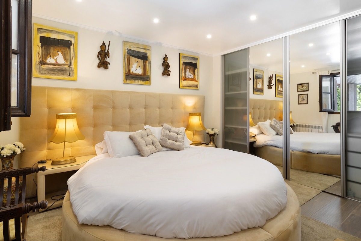 A spacious sleeping area is highlighted by a large round bed with a soft white duvet. Two bedside lamps provide gentle illumination, while framed artwork adorns the walls. A mirrored wardrobe reflects the room, enhancing the sense of space and light.