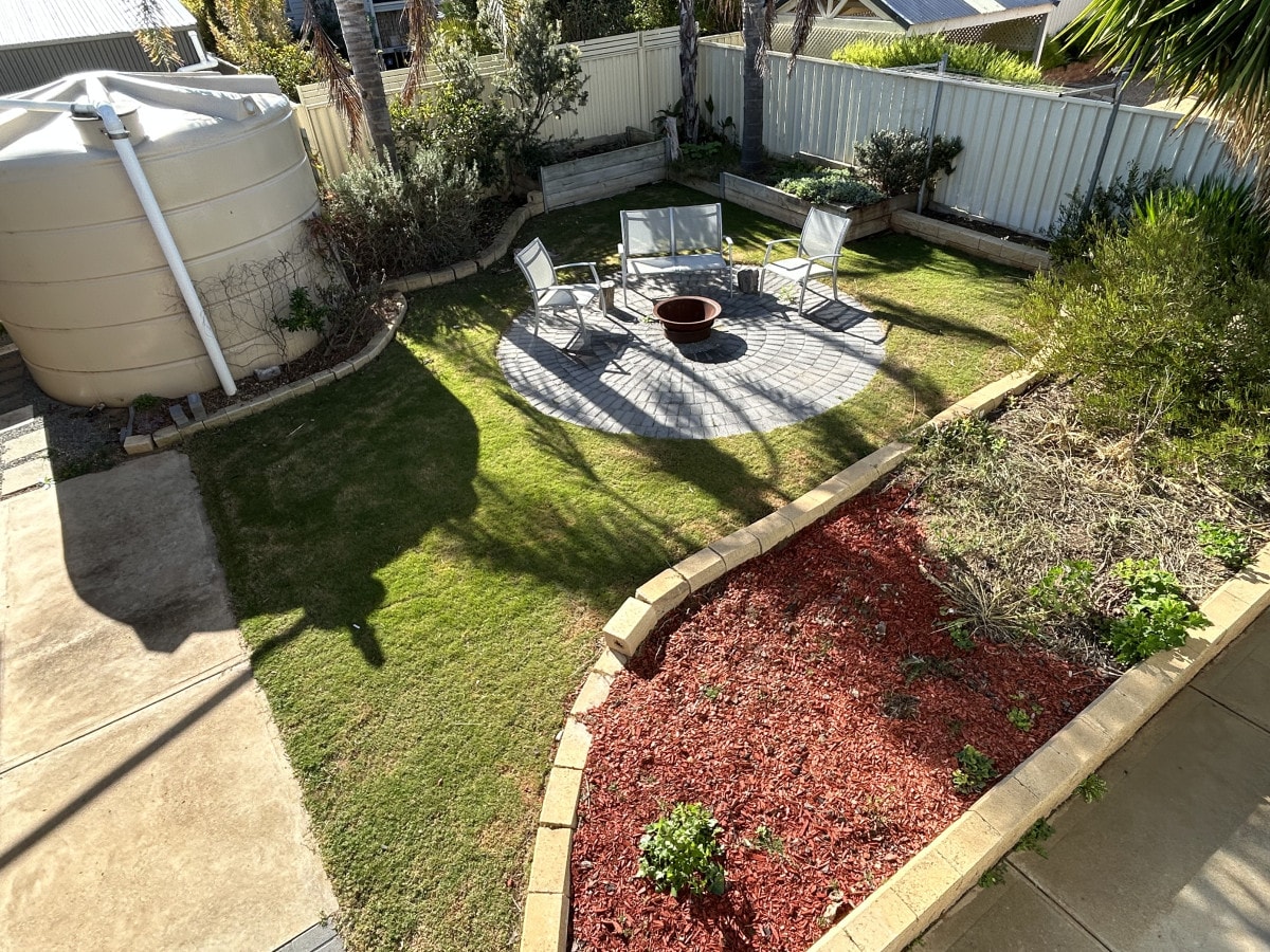 A tranquil outdoor space features a circular stone patio with seating arranged around a firepit. Lush green grass is surrounded by well-maintained garden beds, showcasing a contrast of red mulch and various plants. A large water tank is positioned in the corner of the yard.