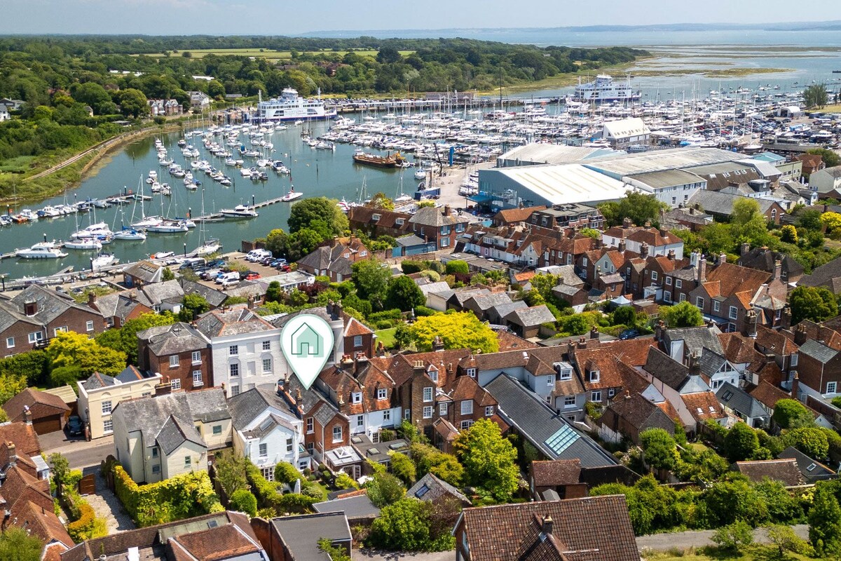 An aerial view captures the charming rooftops of Lymington, with a lush greenery backdrop. The marina features numerous boats moored in the water, alongside the hustle of the nearby residential area. The image conveys the inviting coastal atmosphere of this historic town.