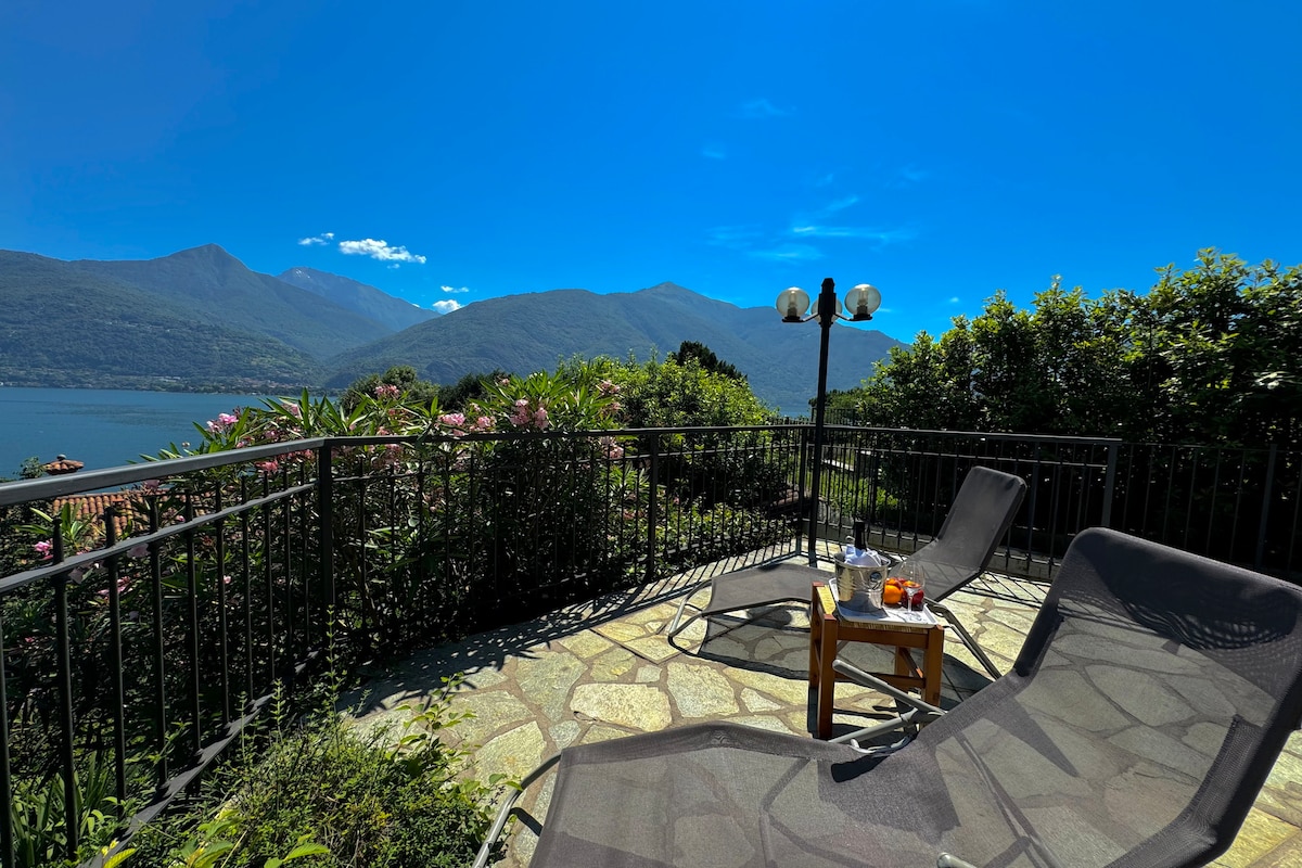 A private terrace features two sun loungers arranged around a small wooden table. Lush greenery surrounds the space, with panoramic views of Lake Como and the surrounding mountains visible under a clear blue sky.