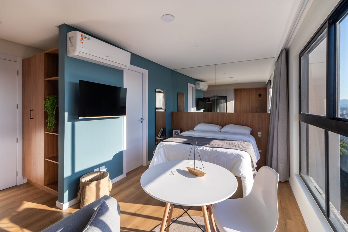 A modern loft interior is displayed, featuring a spacious bed against a mirrored wall, seamlessly reflecting natural light. A round dining table with a triangular centerpiece is positioned nearby, and a cozy seating area is created with a sofa. Large windows offer a view of the surroundings.