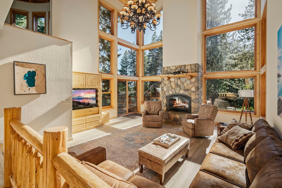 A high-ceiling living room is highlighted by large windows offering views of the surrounding trees. A stone fireplace is centered along one wall, and a comfortable seating arrangement includes oversized sofas and a coffee table. Soft lighting from a chandelier enhances the spacious atmosphere.