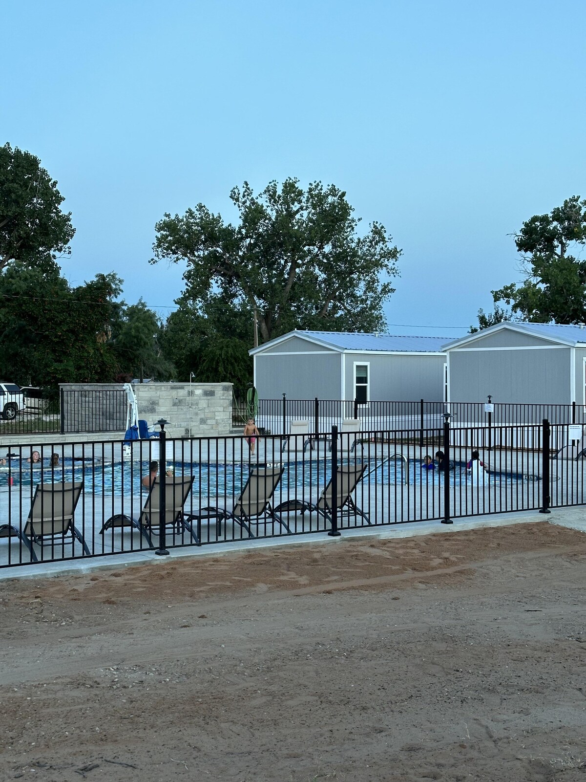 A fenced pool area features several lounge chairs arranged around the water. Green trees provide shade nearby, while modern cabin-like structures are visible in the background, complementing the tranquil outdoor setting.